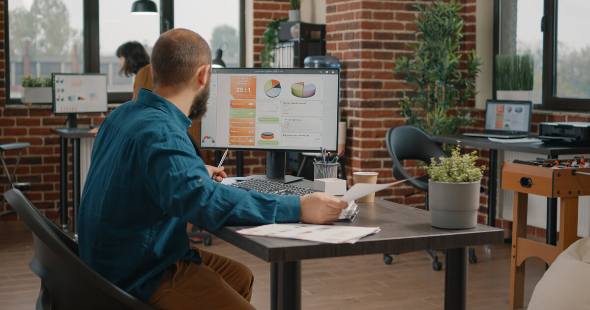 Analyst reviewing data charts on a monitor and printed reports at a desk